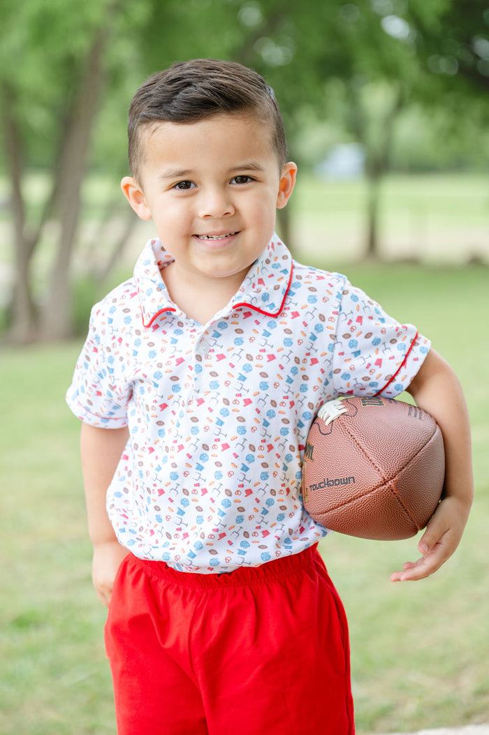 Toddler boy wearing a white Pima cotton polo with football and tailgate print, red piping, and solid red shorts, holding a football