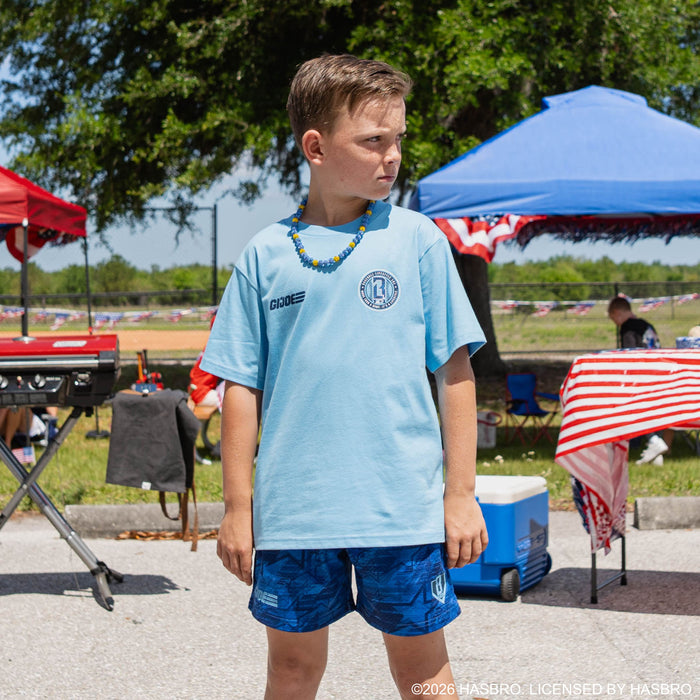 Front-facing youth male model wearing light blue GI Joe tee with blue and yellow beaded necklace at outdoor baseball event