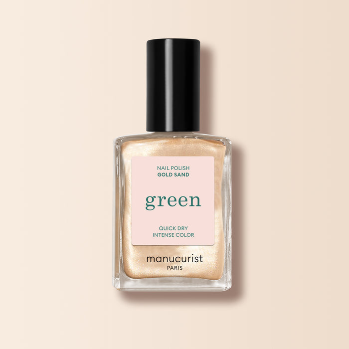 Manucurist Gold Sand nail polish features a shimmering yellow-light gold pearly shade. The bottle has a black cap and a label on pale pink noting “green” and “quick dry intense color.”.