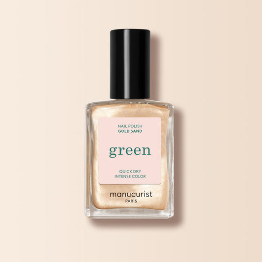 Manucurist Gold Sand nail polish features a shimmering yellow-light gold pearly shade. The bottle has a black cap and a label on pale pink noting “green” and “quick dry intense color.”.