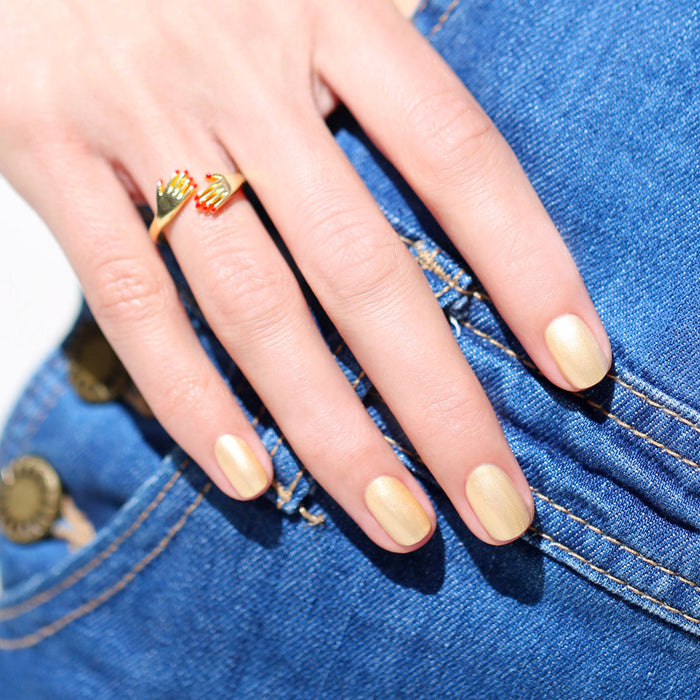 A hand with Manucurist’s Gold Sand iridescent yellow nail polish and a gold double-heart ring rests on blue denim jeans near a pocket with visible stitching and a brass button.