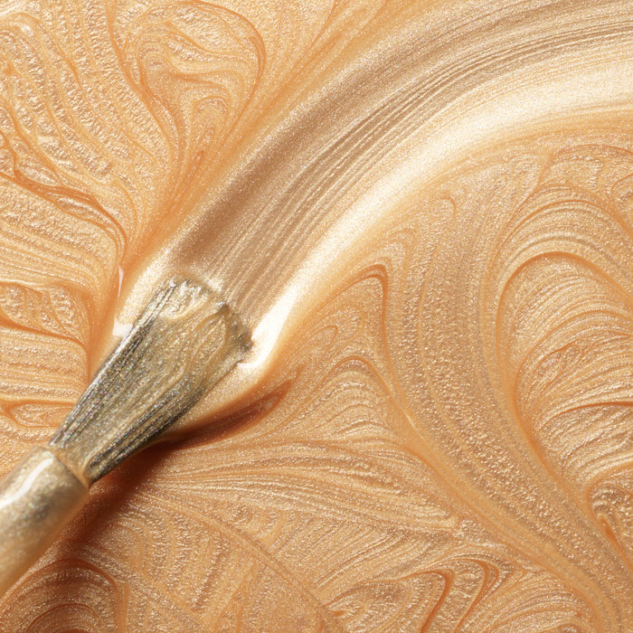 A close-up of a brush spreading shimmering, metallic gold paint with swirling patterns—evoking the brilliance of Manucurist's Gold Sand nail polish.