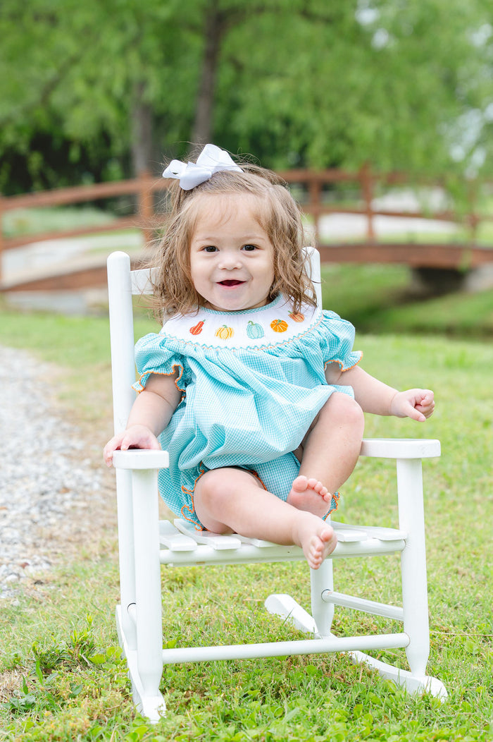 Baby girl sitting on white chair outdoors wearing Smocked Fall Gourds Bubble with white hair bow