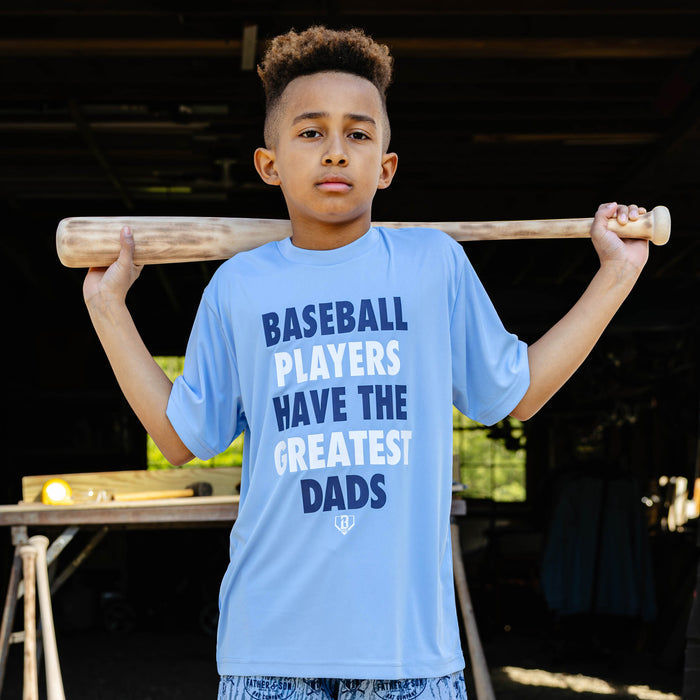 Young boy holding a wooden baseball bat across his shoulders while wearing the columbia blue youth Greatest Dad Athletic Tee with "Baseball Players Have the Greatest Dads" graphic on the front.