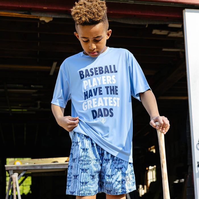 Young boy looking down and pulling the hem of the columbia blue youth Greatest Dad Athletic Tee while paired with Father & Son Bat Company all-over print shorts and holding a baseball bat.