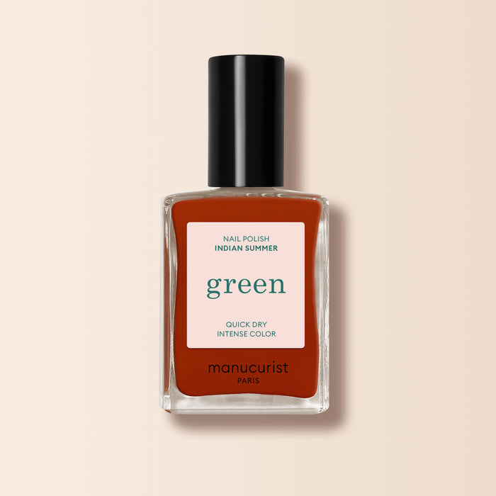 A bottle of Manucurist Indian Summer nail polish in brick red, from the Green™ range, comes in a rectangular glass bottle with a black cap and a label highlighting green, quick dry, and intense color on a light beige background.