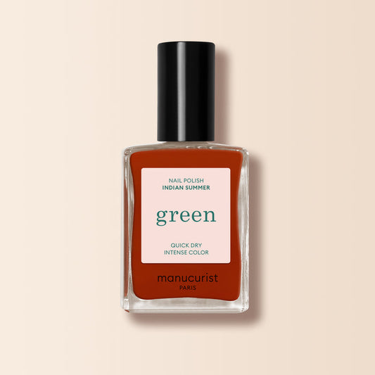 A bottle of Manucurist Indian Summer nail polish in brick red, from the Green™ range, comes in a rectangular glass bottle with a black cap and a label highlighting green, quick dry, and intense color on a light beige background.