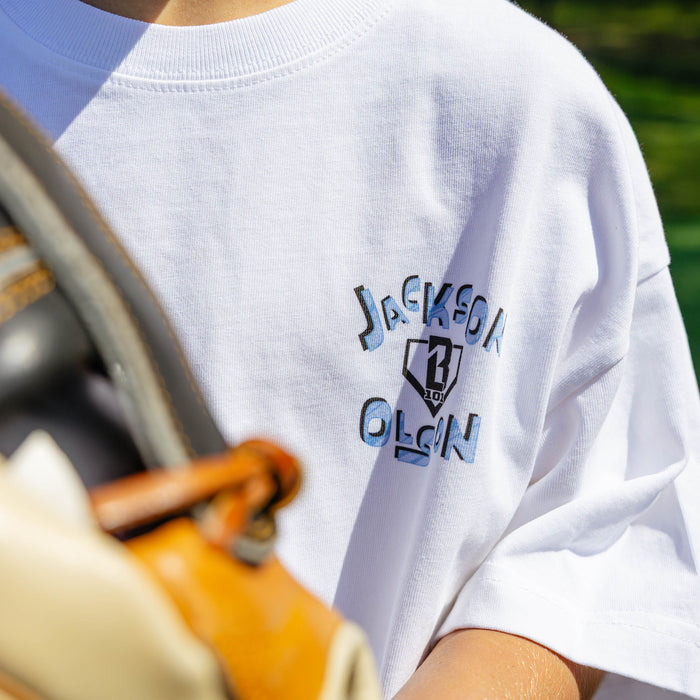 Close-up of the Jackson Olson x BL101 chest graphic on the youth white tee, featuring blue and black lettering around the BL101 shield logo