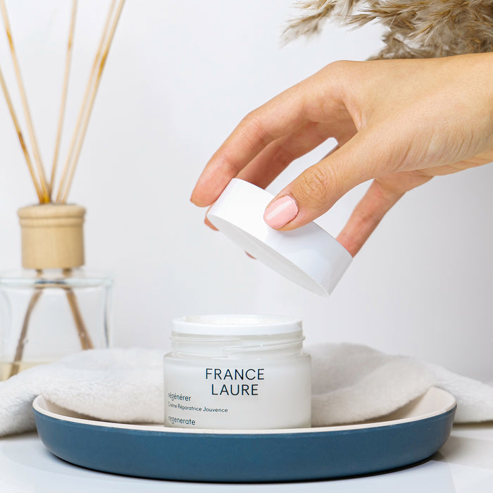 Hand opening France Laure Regenerate Repair Youth Cream jar, showcasing luxurious skincare for all skin types with elegant white packaging.