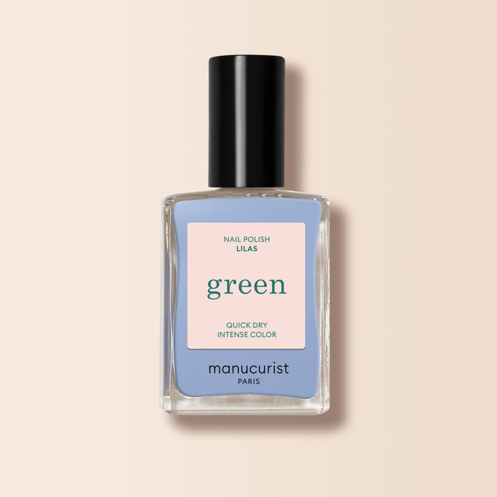 A bottle of Manucurist Lilas nail polish with a black cap and “Nail Polish Lilas, Quick Dry, Intense Color” label sits on a light beige background next to pastel blue polish.