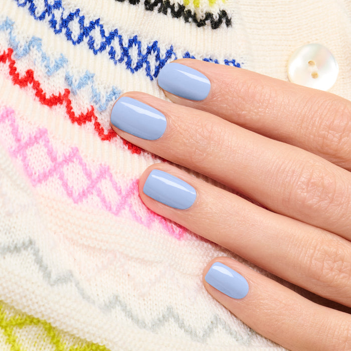 A hand with short, neatly manicured nails painted in Manucurist’s Lilas pastel blue nail polish rests on a white knitted sweater featuring colorful zigzag patterns and a white button.