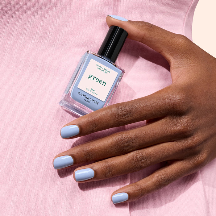 A hand with pastel blue nail polish holds a bottle of Manucurist Lilas nail polish, set against a pale pink fabric background.