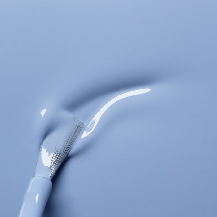 A close-up of the Manucurist Lilas white brush applying pastel blue nail polish, creating a smooth, glossy streak on a matching blue surface.