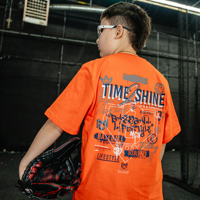 Child wearing an orange t-shirt with graphics and text, holding a baseball glove indoors.