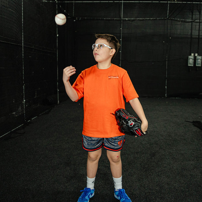Child in an indoor batting cage wearing an orange shirt and blue shoes, preparing to catch a ball.