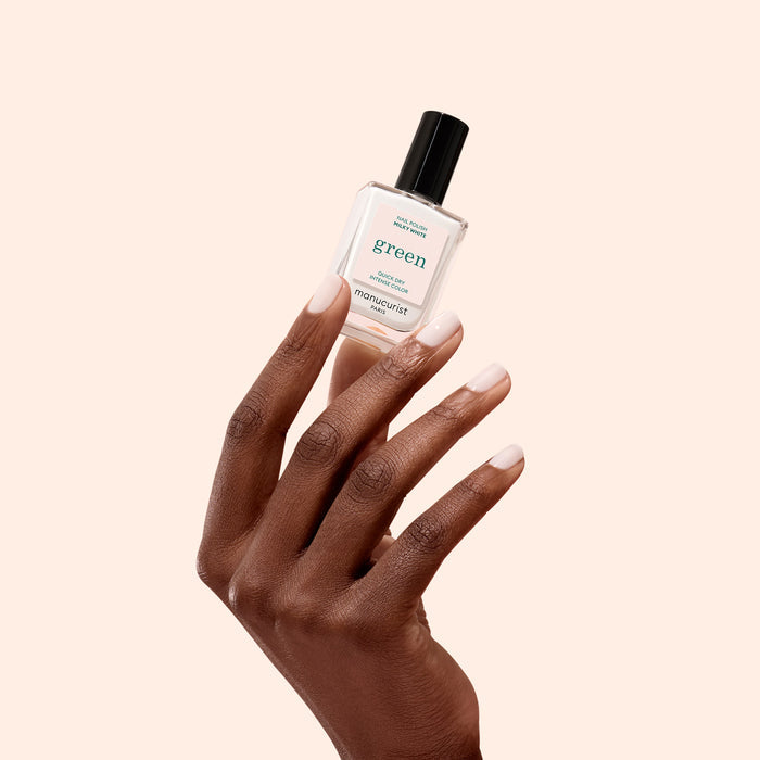 A hand with neatly manicured nails holds a bottle of Milky White nail polish from Manucurist’s Green™ range against a light beige background.