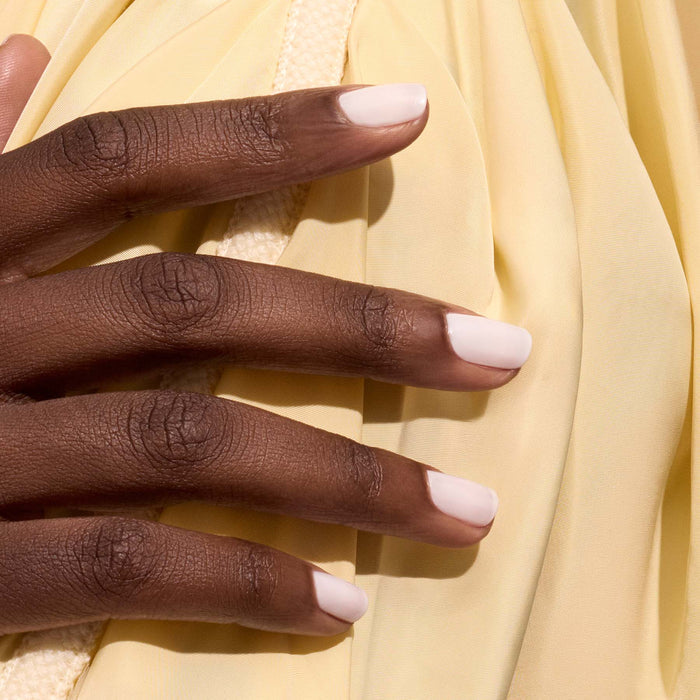 A close-up of a hand with medium-dark skin and neatly manicured nails painted in Milky White by Manucurist, resting on soft, pale yellow fabric.