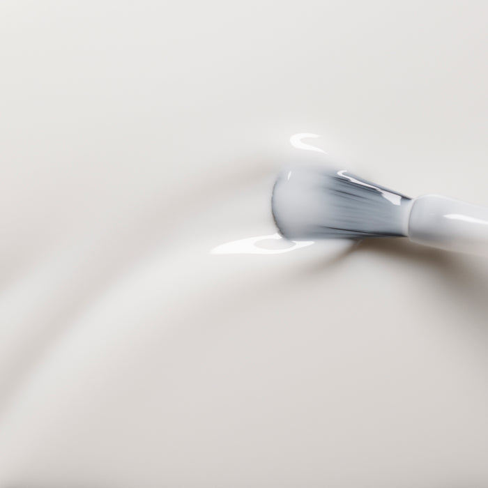 A close-up of a soft brush spreading Manucurist’s Green™ Milky White nail polish—a smooth, creamy white formula—on a surface, forming gentle ripples and glossy highlights.
