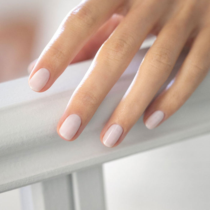 A close-up of a hand resting on a white wooden surface, showing short, neatly manicured nails painted in Milky White by Manucurist for a soft, pale pink glossy finish.