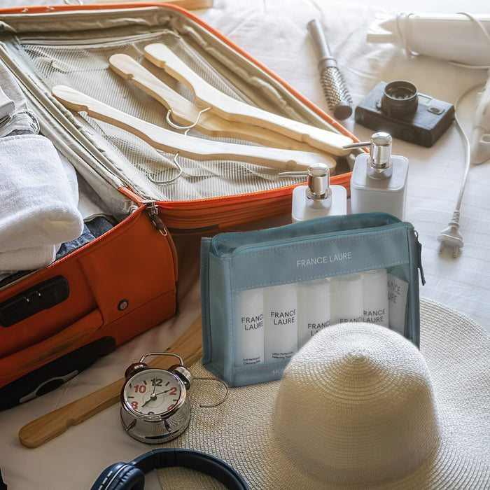 France Laure travel skincare set with cleansing milk and toner in a blue pouch beside an open suitcase, hat, and camera for vacation prep