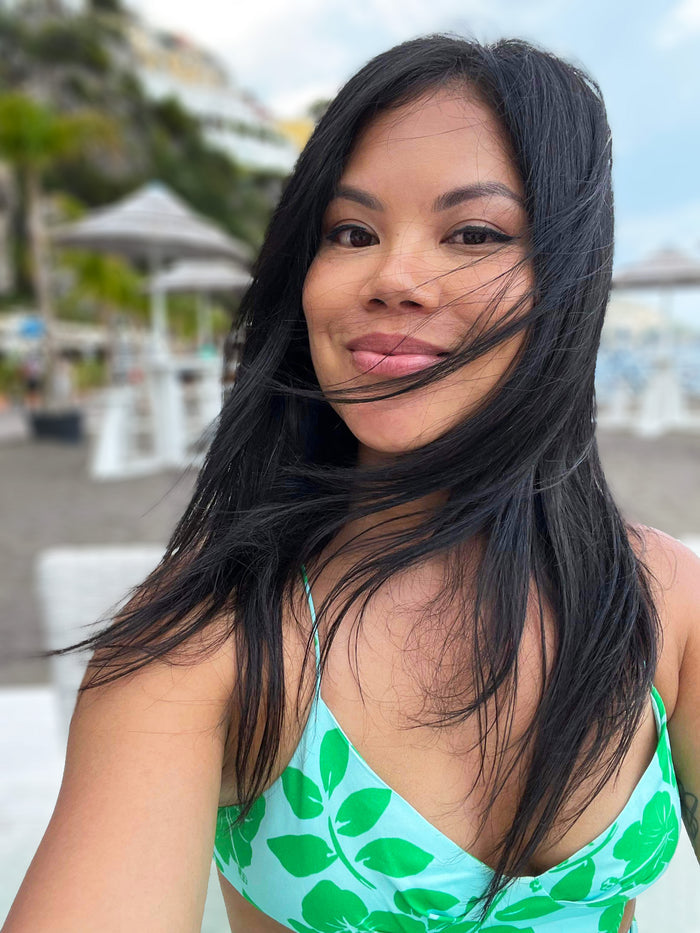 Woman with long dark hair in green leaf print bikini top, smiling at beach resort with palm trees and cabanas