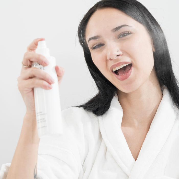 Woman in white robe joyfully misting face with skincare spray, showcasing refreshing facial mist application for all skin types