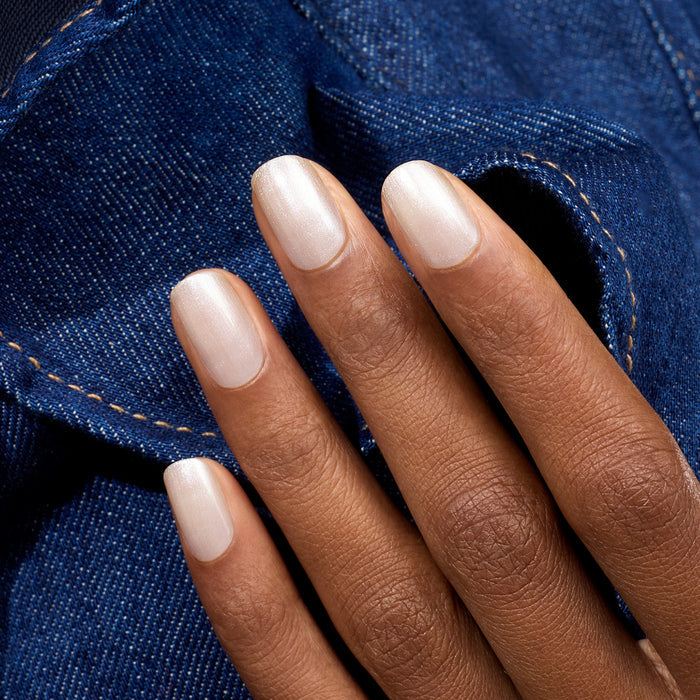 A hand with medium brown skin and neatly manicured nails painted in Manucurist’s Moon, a pearly white polish, rests against blue denim fabric with visible stitching.