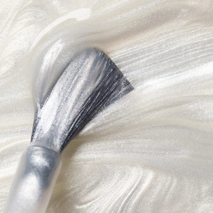 A close-up of a brush applying Manucurist's Moon shimmery pearly white nail polish, creating smooth, glossy streaks on a surface.