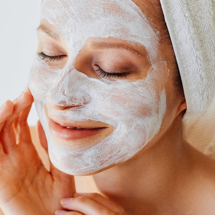Woman applying creamy white face mask with towel wrapped around head, enjoying relaxing skincare routine for all skin types