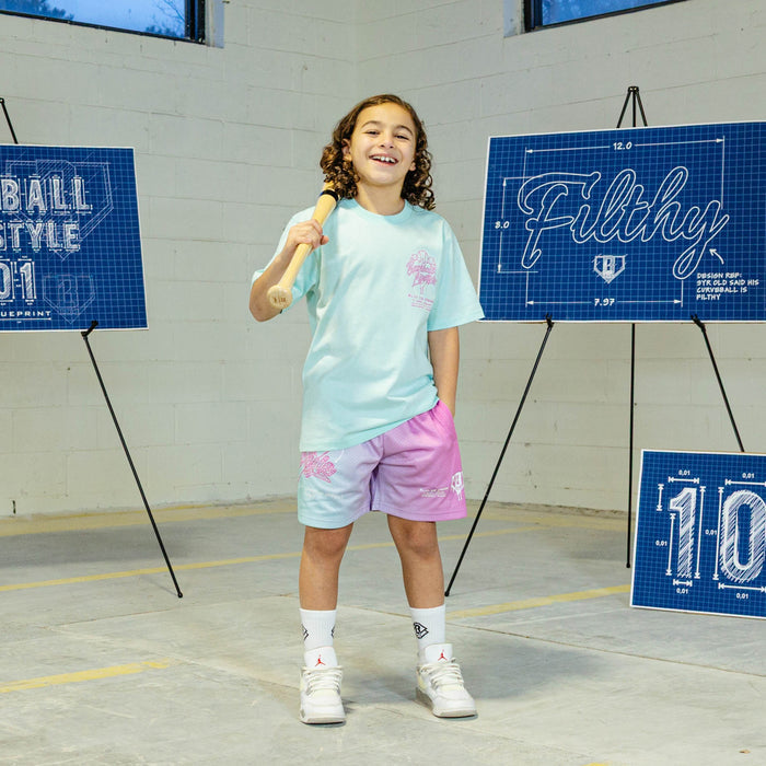 Child holding a baseball bat in an indoor sports facility wearing light blue t-shirt with pink ice cream graphic and "Baseball Lifestyle 101" logo