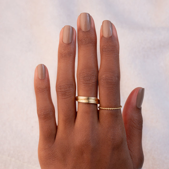 A hand with neatly manicured Orme nails from Manucurist wears two gold rings—a smooth band on the middle finger and a twisted rope ring on the ring finger—set against a soft, light background.
