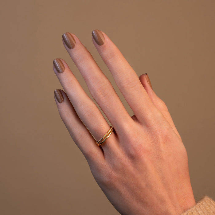 A hand with neatly manicured nails painted in Orme, a light brown polish by Manucurist, is shown against a beige background. The hand wears a simple gold ring on the middle finger.