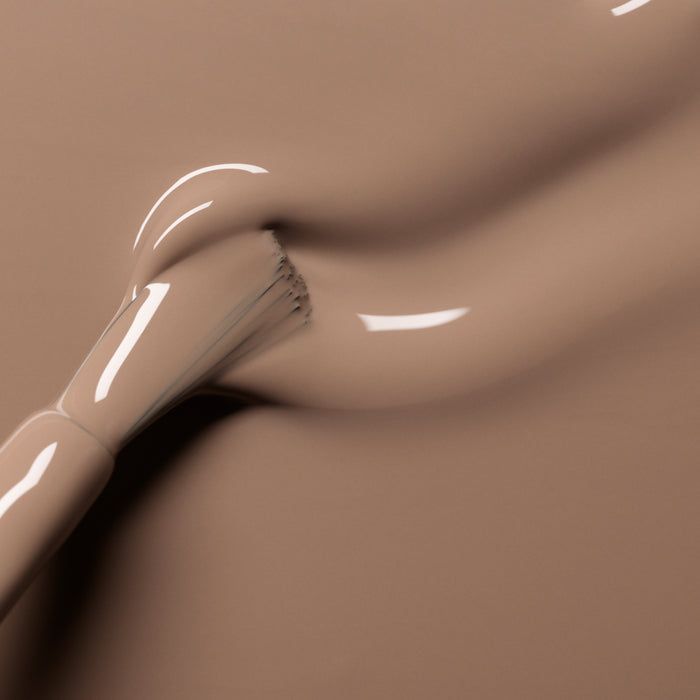 A close-up of Manucurist’s Orme makeup brush spreading smooth, café au lait liquid foundation on a matching background, forming soft curves and glossy highlights.