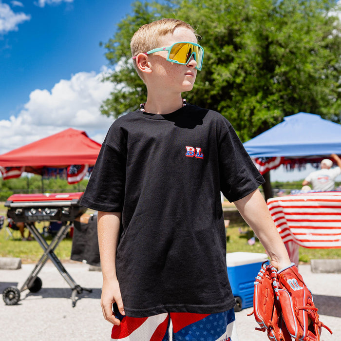 Front-facing youth male model wearing black Patriot Pitch tee with red white and blue shorts holding baseball glove with teal shield sunglasses at outdoor baseball event