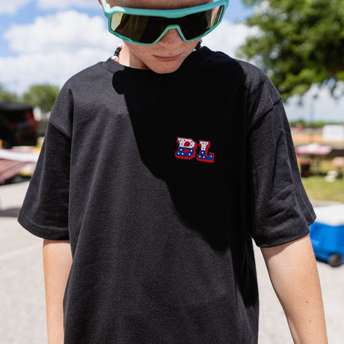Close-up of youth male model wearing black Patriot Pitch tee with teal shield sunglasses showing red white and blue BL logo on chest at outdoor event