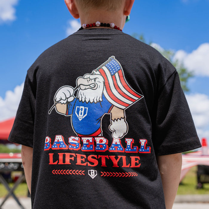 Black: Back view of youth male model showing Baseball Lifestyle eagle graphic on black tee at outdoor baseball event