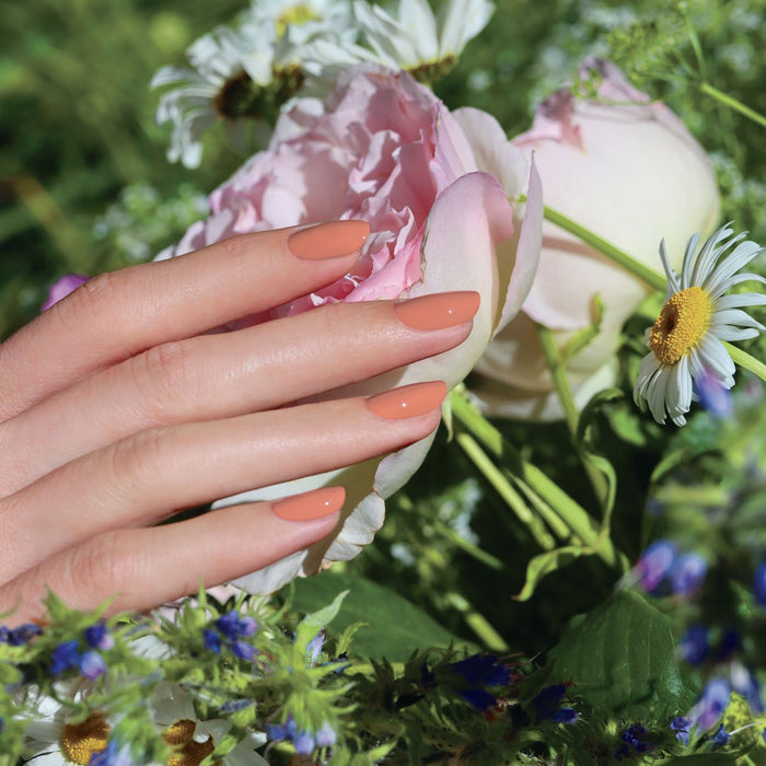 A hand with neatly manicured nails in Manucurist's Peach shade gently rests on a light pink peony, surrounded by daisies and green foliage in a garden setting.