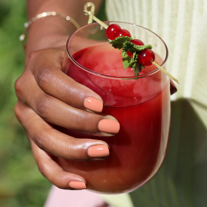 A hand with Manucurist Peach nail polish holds a glass of red juice with ice, mint, and red currants, set against a blurred green background.