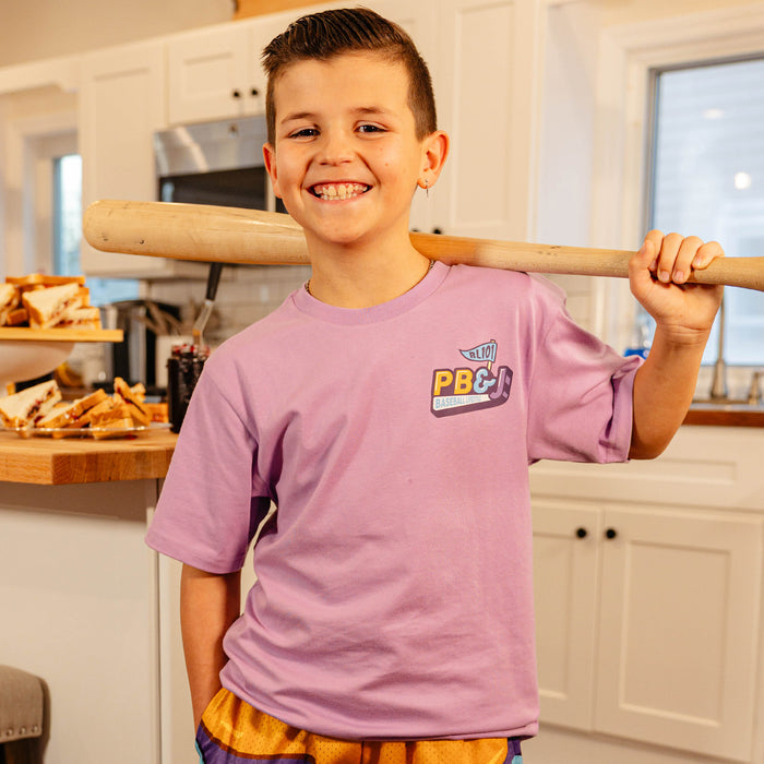 Young boy smiling in a kitchen wearing the BL101 youth PB&J Baseball Lifestyle purple tee with a baseball bat over his shoulder.