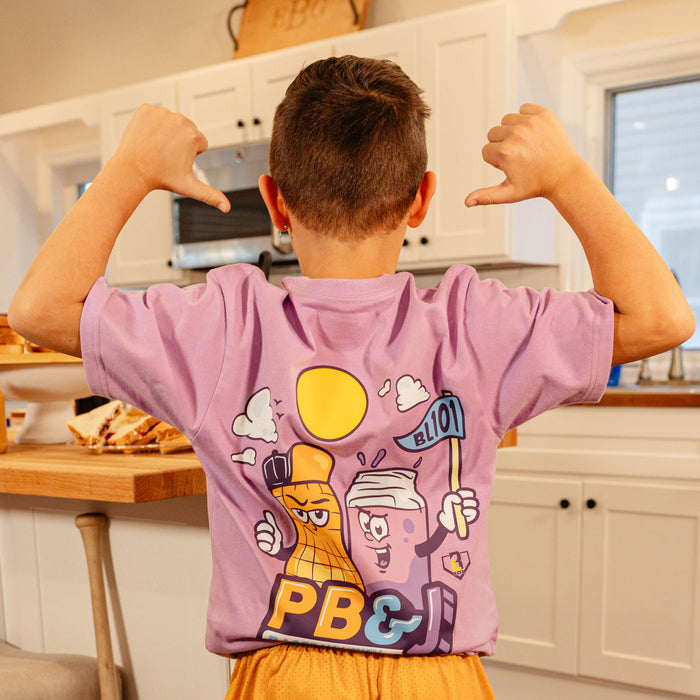 Back view of a young boy pointing to the cartoon peanut and jelly jar mascot graphic on the BL101 youth PB&J Baseball Lifestyle purple tee.