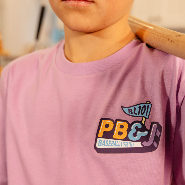 Close-up of the PB&J Baseball Lifestyle chest logo on the youth purple tee, with a baseball bat resting on the shoulder.
