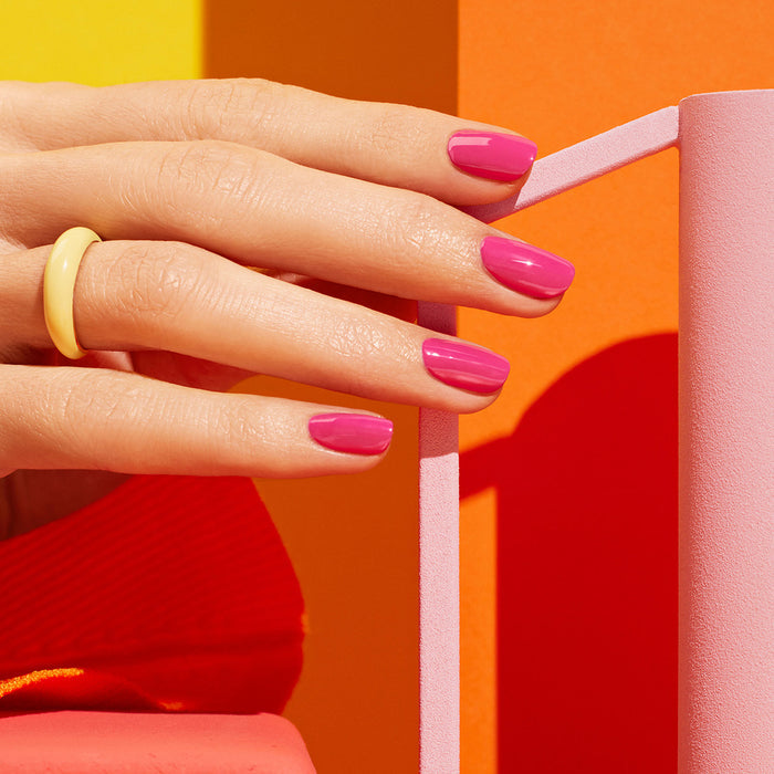 A hand with glossy pink "Pétula" nail polish by Manucurist and a chunky yellow ring rests against bold pink and orange shapes on an orange background, while a red sleeve highlights the vibrant color play with subtle blue undertones.