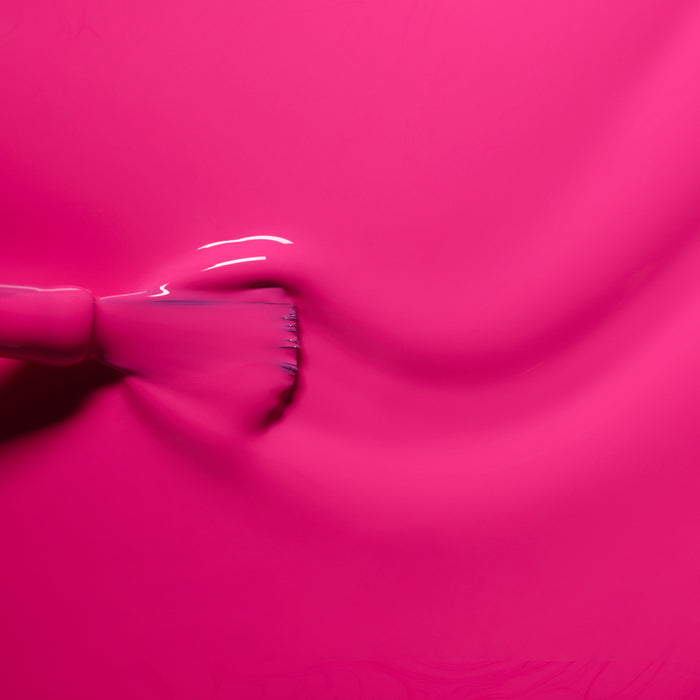 A close-up of a brush spreading Manucurist's Pétula, a bright pink nail polish, on a smooth surface, revealing glossy finish, blue undertones, and visible brush marks.