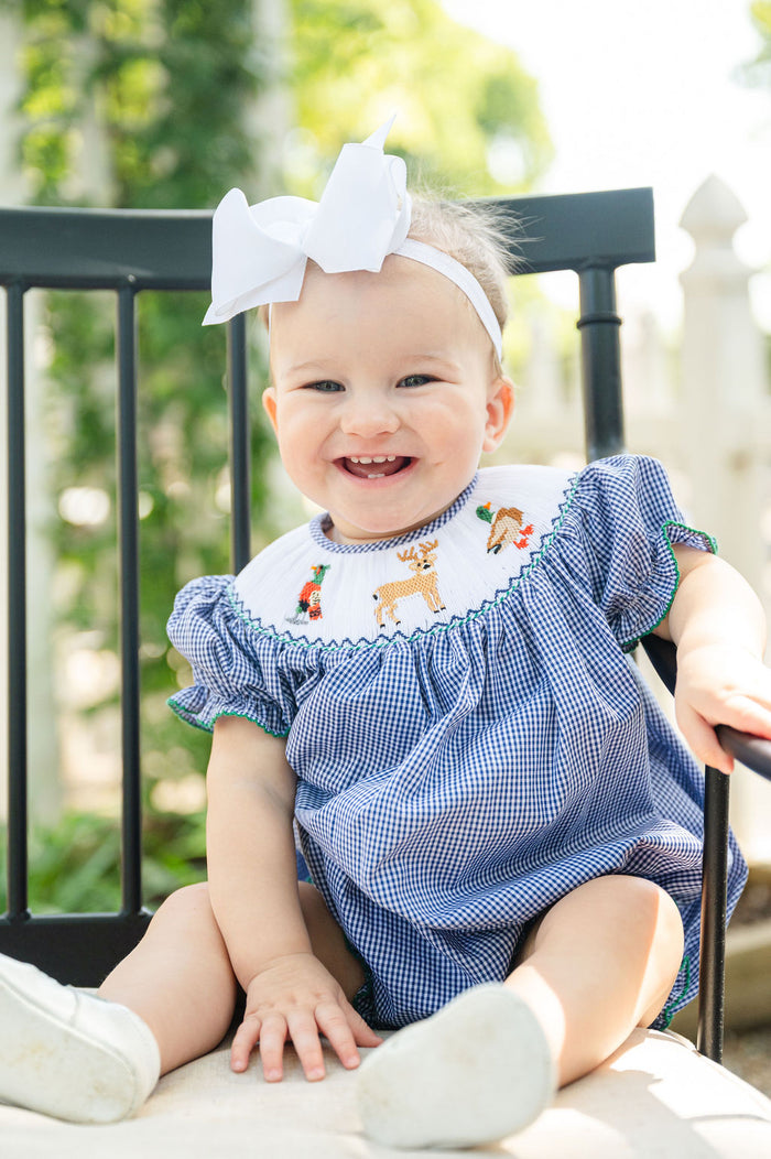 Baby girl wearing Smocked Hunting Season Bubble by Ruth and Ralph, blue gingham with white bow, sitting outdoors