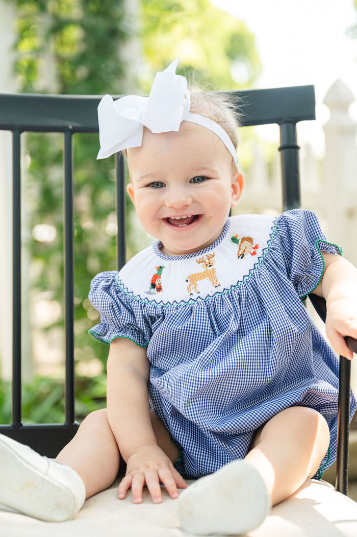 Close-up of baby girl smiling in Smocked Hunting Season Bubble by Ruth and Ralph, sitting outdoors with white bow