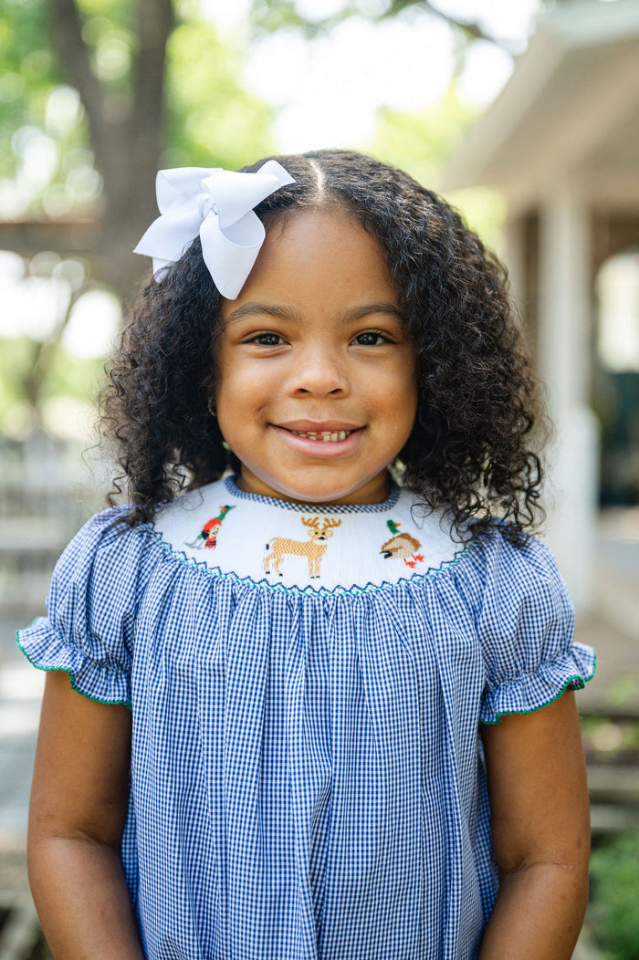 Girl smiling in Smocked Hunting Season Dress by Ruth and Ralph, blue gingham with white bow, outdoors