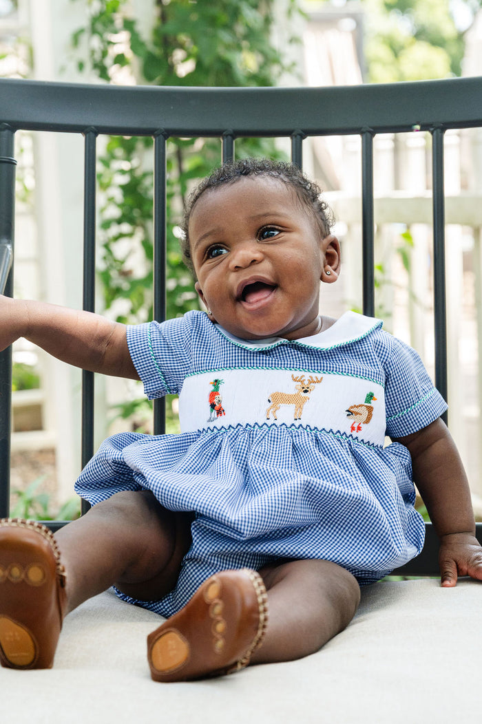 Baby wearing Smocked Hunting Bubble by Ruth and Ralph, blue gingham with smocked deer and duck embroidery, sitting outdoors