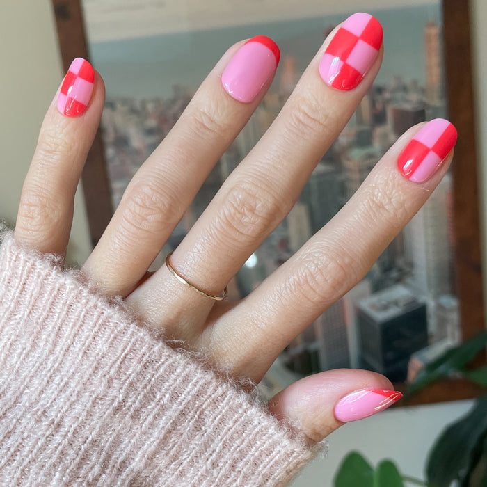 Pink Checker Party | Short | Round - Tab Press-On showing hand with pink and red checker and solid design false nails indoors