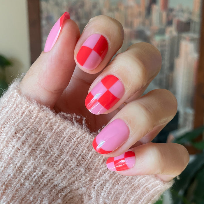 Pink Checker Party | Short | Round - Tab Press-On showing hand with pink and red checker false nails indoors with cozy sweater sleeve