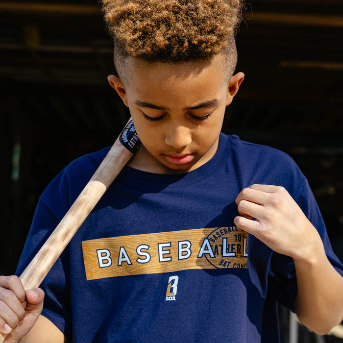 Close-up of a young boy looking down and pulling the front of the navy youth Radar Tee to show the wood grain baseball graphic and Father & Son Bat Company stamp print.
